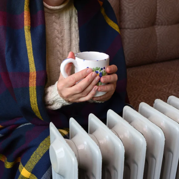 A woman enjoys a warm mug of coffee while sitting in front of a radiator, illustrating solutions to keep hands warm.