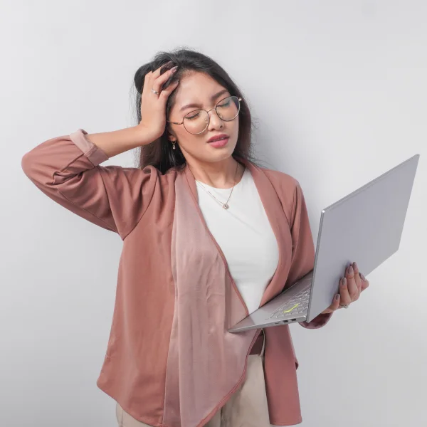 Asian woman with glasses and a laptop, visibly stressed, holding her head while considering the effects of Raynaud's Syndrome.