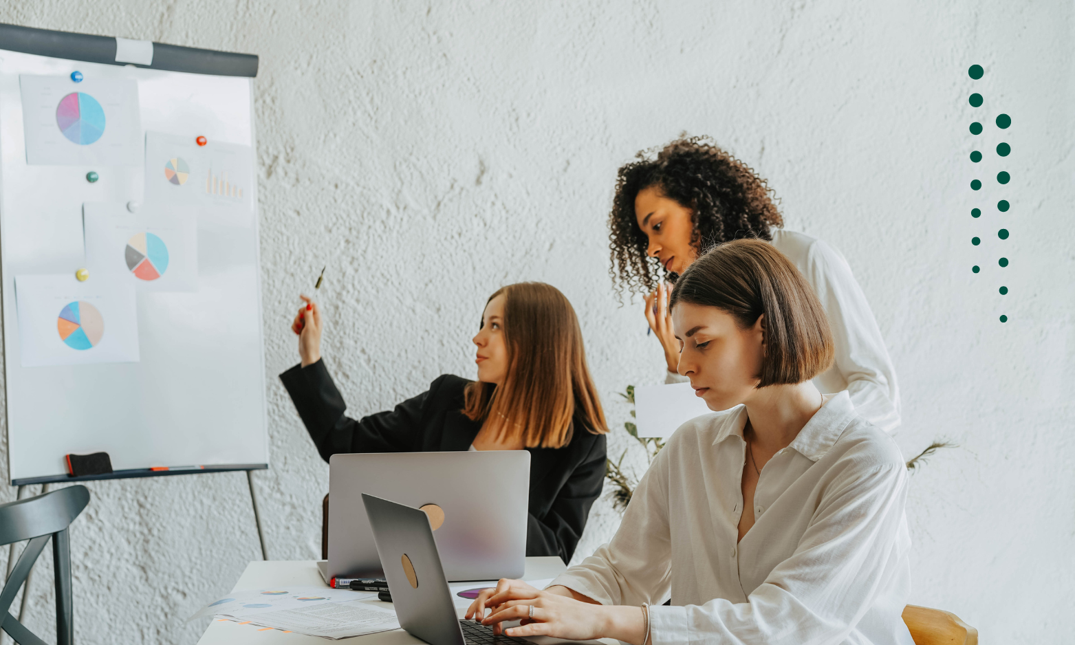 Trois femmes collaborent dans un espace de bureau minimaliste avec un mur blanc texturé en arrière-plan. À gauche, une femme aux cheveux bruns longs, portant un blazer noir, pointe vers un tableau blanc affichant des graphiques en secteurs colorés et des diagrammes à barres. Au centre, une femme aux cheveux bouclés s'incline légèrement, participant activement à la discussion. À droite, une femme avec une coupe carrée brune et une chemise blanche se concentre sur son ordinateur portable tout en travaillant à la table, qui est couverte de papiers et d'outils d'écriture. La scène illustre le travail d'équipe et la productivité dans un environnement professionnel.