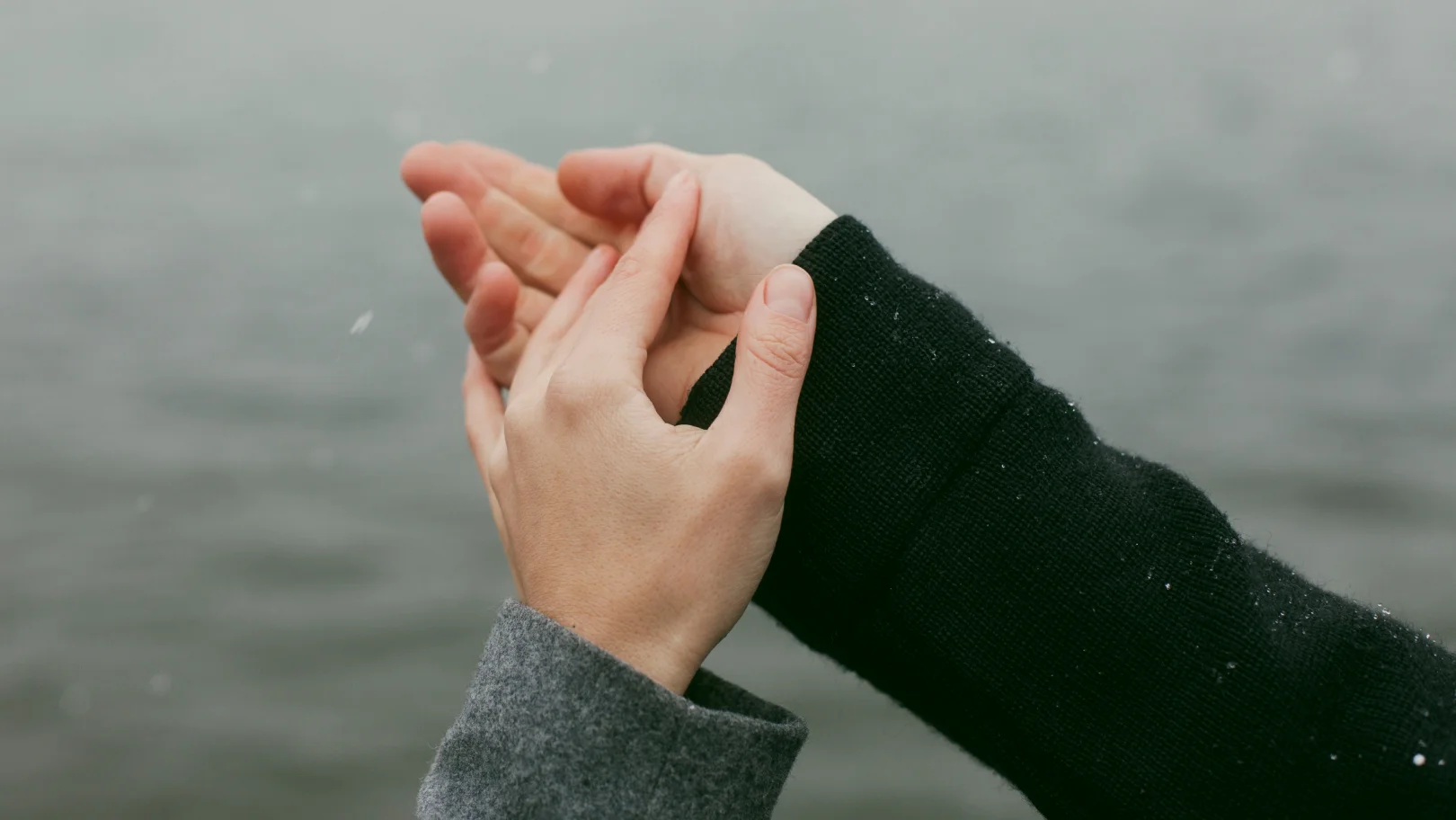 A couple holding hands by a body of water, both appearing to have cold hands as they enjoy the serene view.