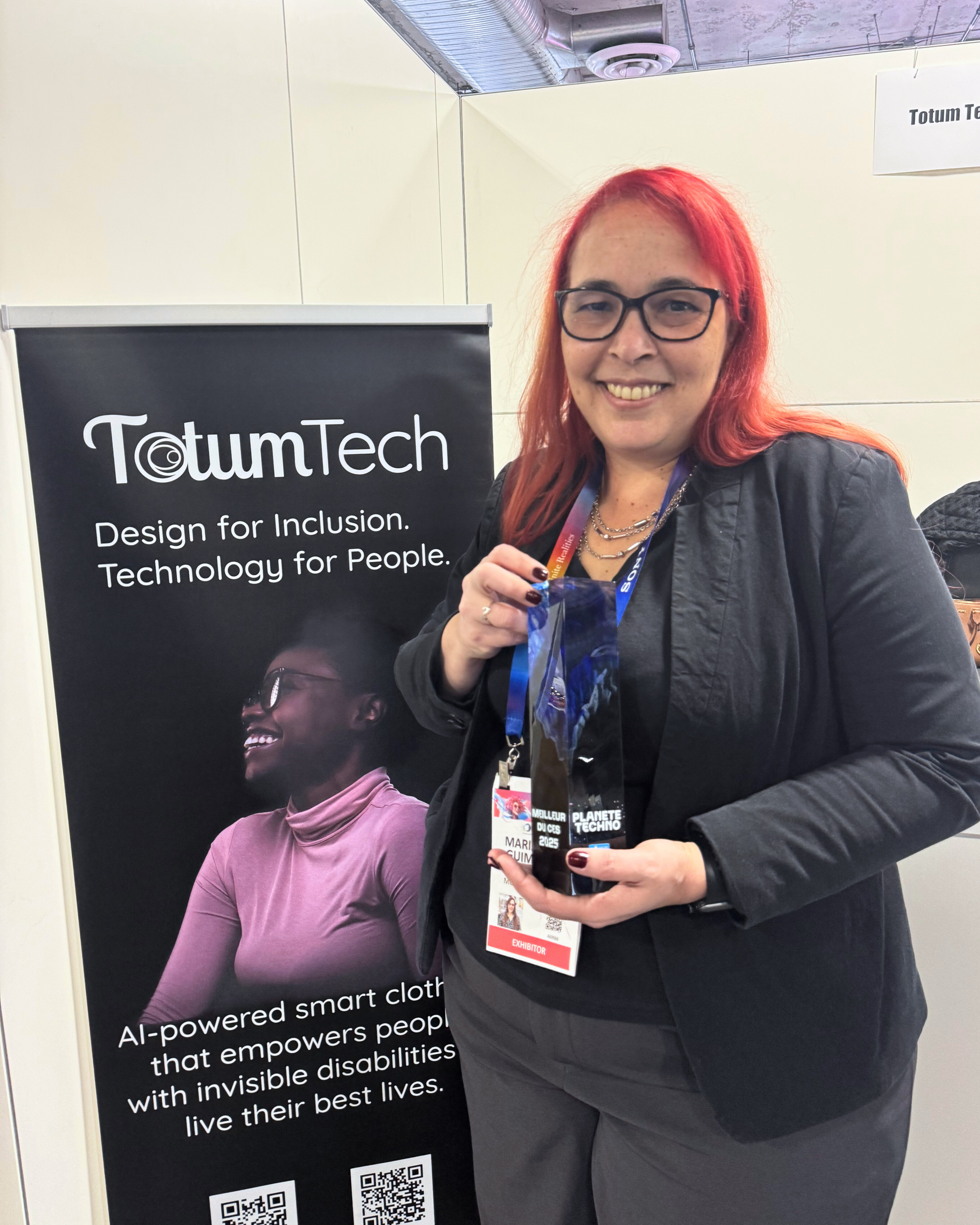 Maria Julia Guimaraes, PDG de Totum Tech, souriante en tenant le prix “Meilleur du CES 2025” de Planète Techno. Elle se tient devant une bannière Totum Tech mettant en avant la mission de vêtements intelligents inclusifs propulsés par l’IA.